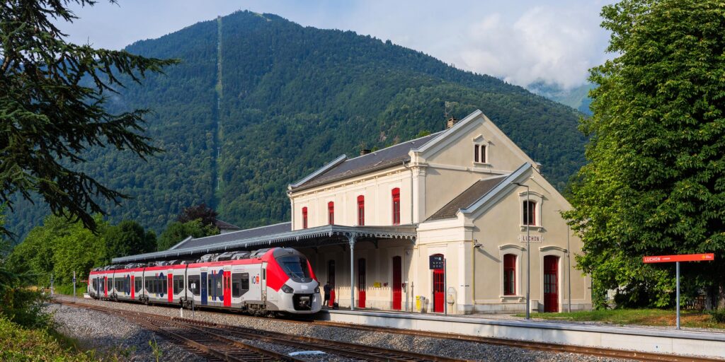 Gare de Bagnères-de-Luchon rénovée avec train LiO à quai, vue sur la montagne de Superbagnères et les lignes des télécabines.