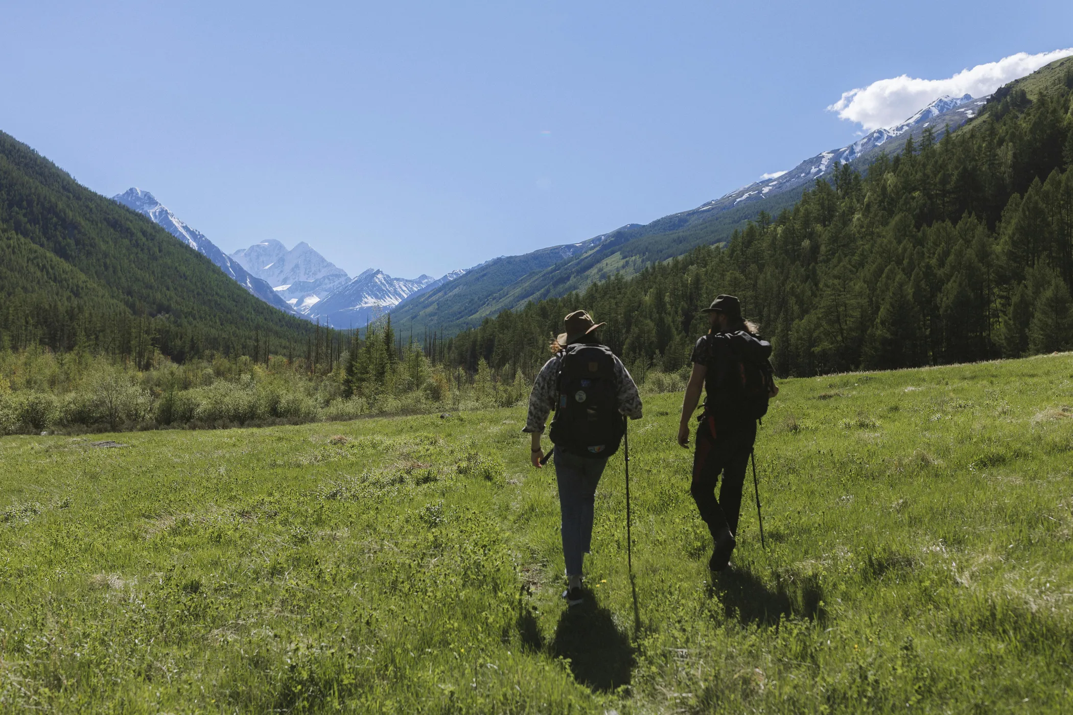 Deux randonneurs avec sacs à dos et bâtons marchant dans une prairie verdoyante des Pyrénées face aux sommets enneigés du Luchonnais.