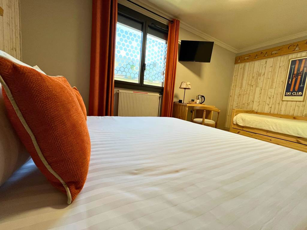 Vue depuis le lit de la chambre n°6 montrant la télévision écran plat murale, le coin bureau et la fenêtre avec ses rideaux orange.