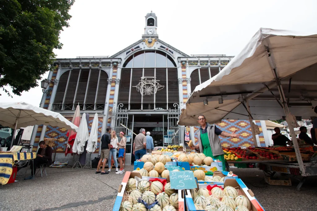 Marché de plein air à Luchon avec étal de primeur au premier plan et la halle gourmande historique en arrière-plan sur la place du marché.