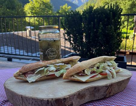 Deux sandwichs baguettes garnis frais et gourmands sur une planche en bois, avec une vue ensoleillée sur les montagnes des Pyrénées depuis le balcon de l'Hôtel La Rencluse à Luchon. Parfait pour une randonnée.