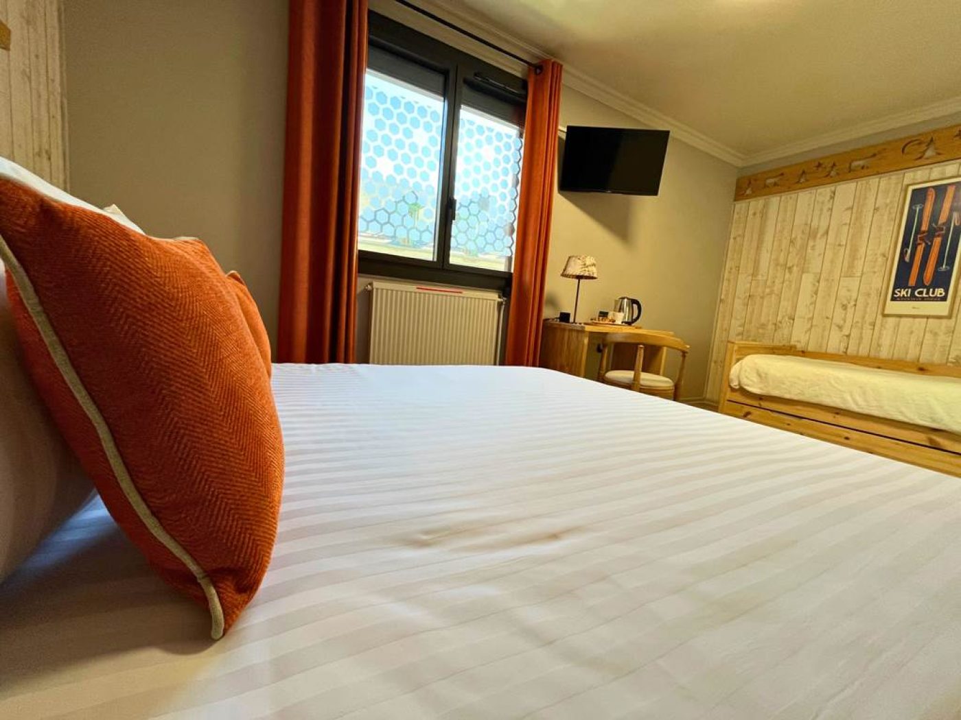 Vue depuis le lit de la chambre n°6 montrant la télévision écran plat murale, le coin bureau et la fenêtre avec ses rideaux orange.