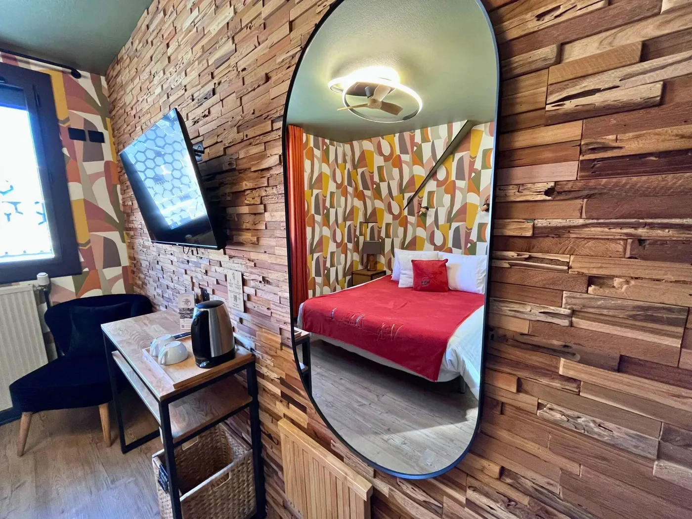 Détail du mur en parement de bois avec miroir organique reflétant le lit rouge de la chambre 10.