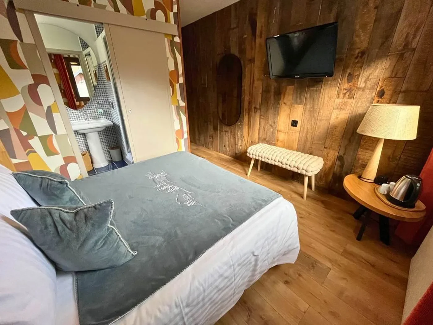 Vue depuis la chambre vers la salle de bain privative ouverte, montrant un lavabo colonne blanc, un miroir rond et une décoration soignée.