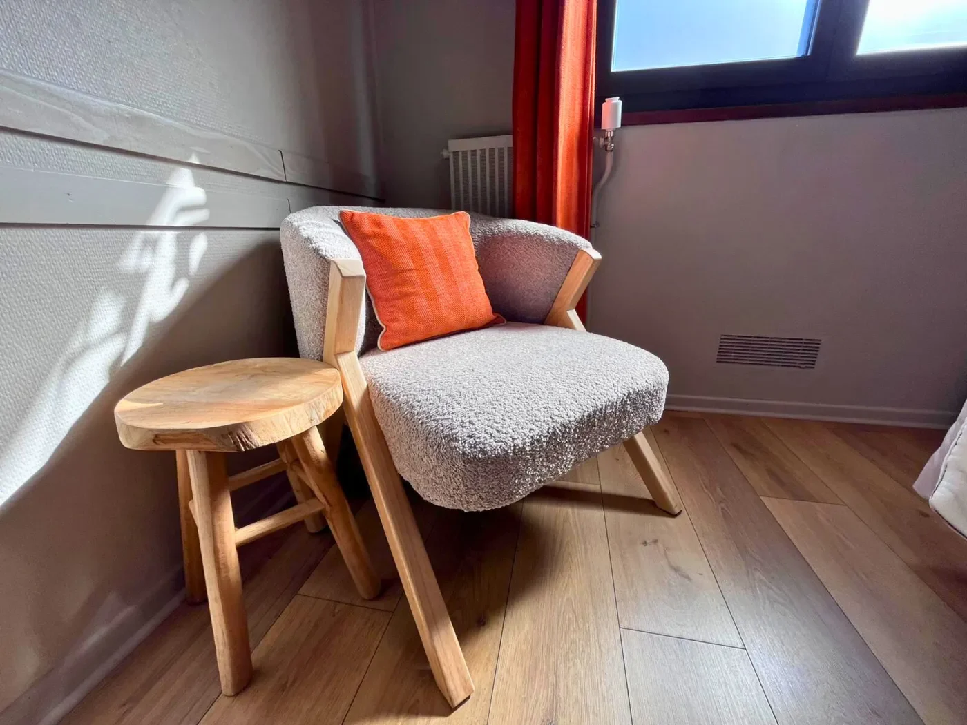 Coin détente avec fauteuil en bouclé gris, coussin orange vif et tabouret en bois brut dans une chambre de l'Hôtel La Rencluse.