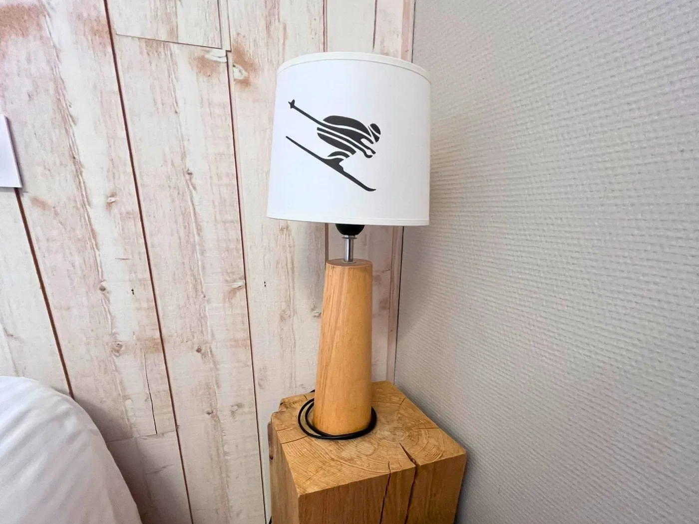 Détail d'une lampe de chevet design en bois avec un skieur vintage illustré sur l'abat-jour, posée sur un billot de bois brut.