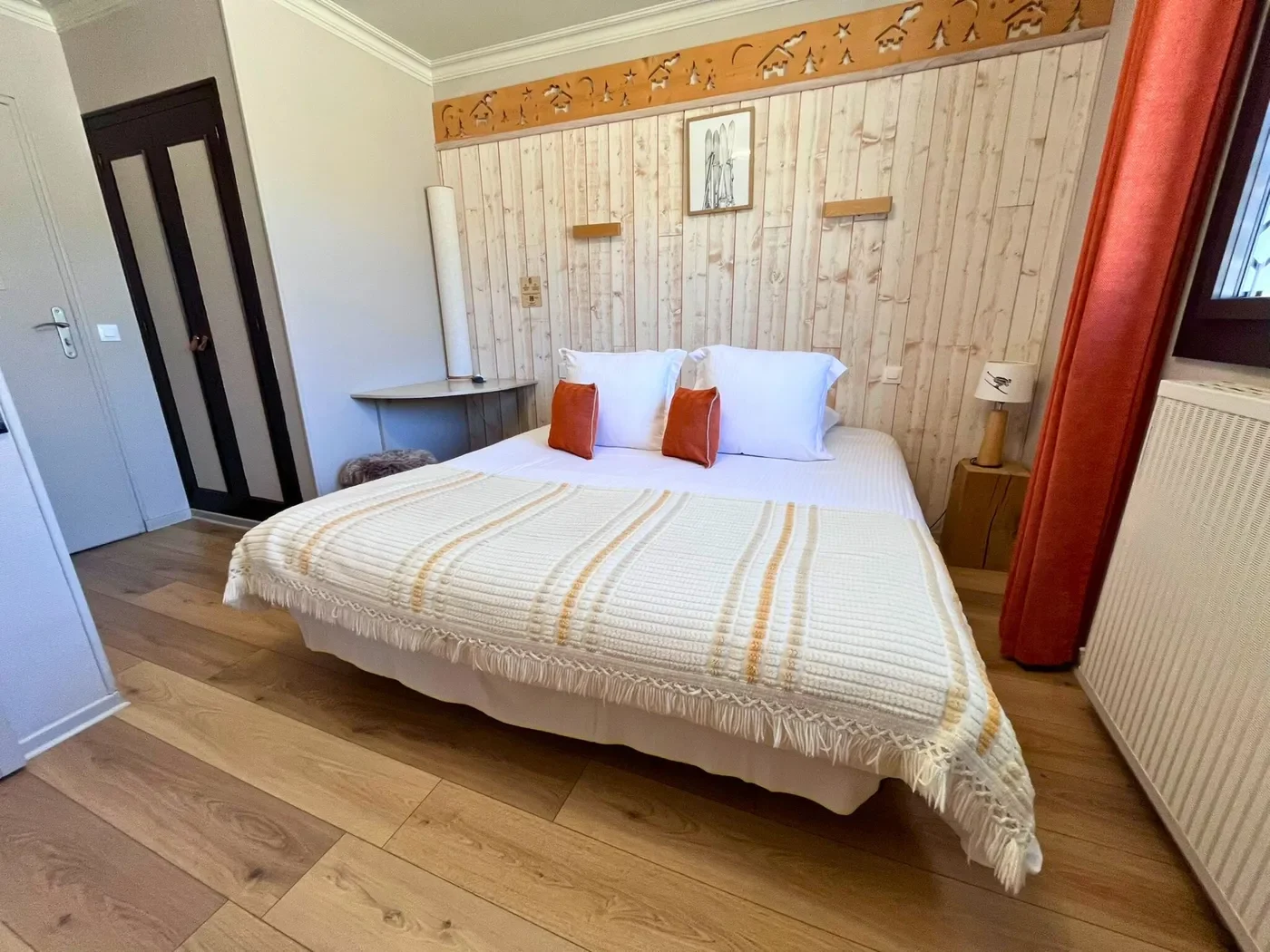 Grand lit double avec plaid rayé, coussins orange et tête de lit en bois sculpté d'une frise montagnarde en chambre 14.