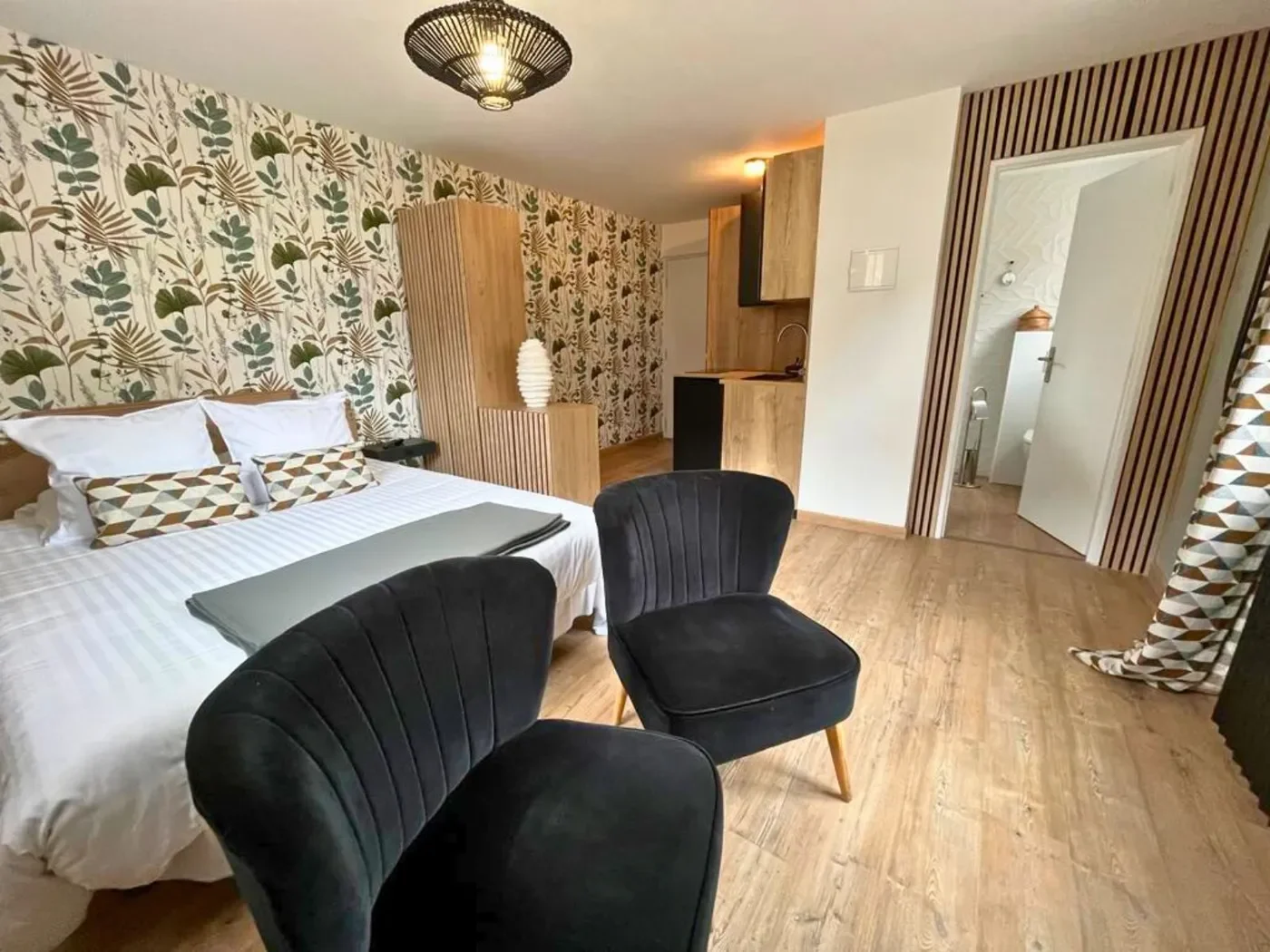Vue d'ensemble du studio annexe montrant l'espace cuisine moderne en bois, le coin repas avec fauteuils noirs et le lit queen-size.