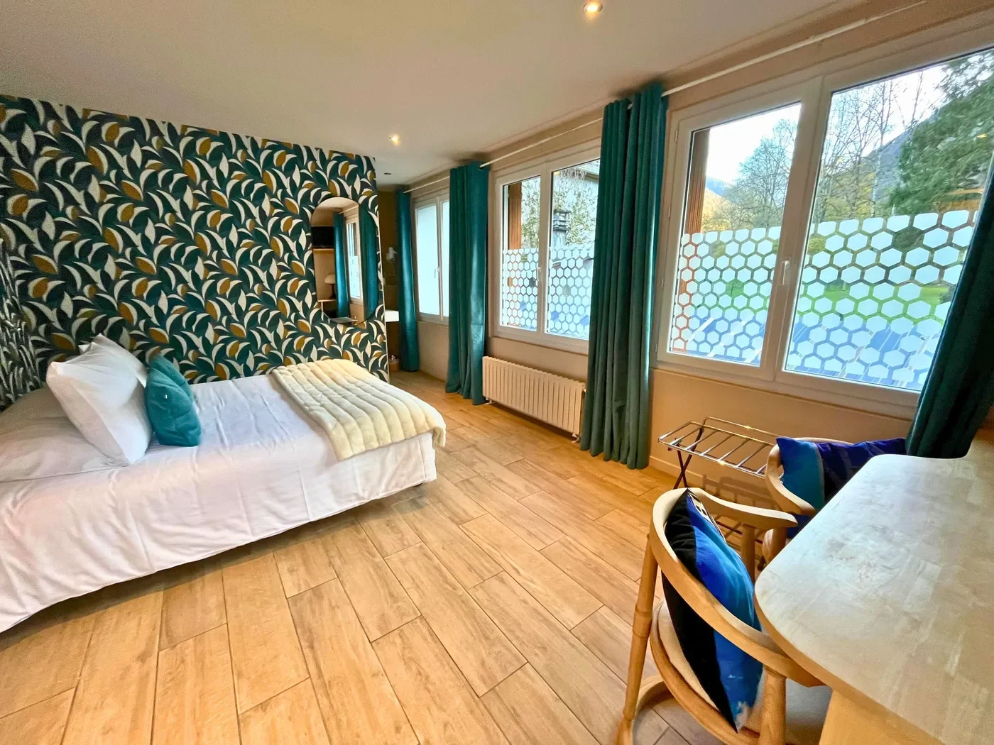 Perspective de la chambre 32 montrant les grandes baies vitrées lumineuses, les rideaux turquoise et le lit double.