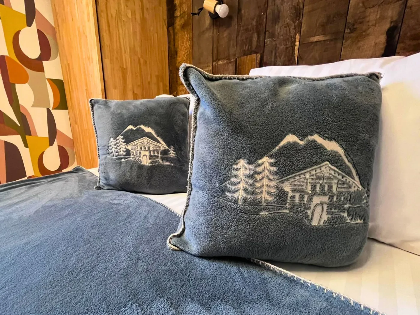 Détail de la literie de la chambre n°2 à l'Hôtel La Rencluse : oreillers en velours gris avec broderies chalets et montagnes sur fond de mur en bois.