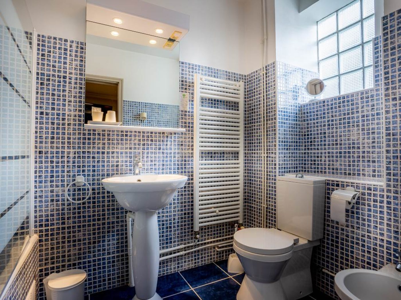 Salle de bain de la chambre 14 avec mosaïques bleues, lavabo colonne blanc et grand miroir éclairé.