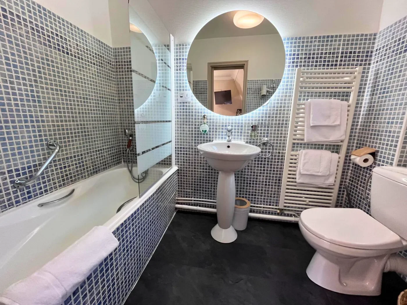 Salle de bain moderne en mosaïque bleue avec miroir rond rétro-éclairé, baignoire et douchette géothermale.